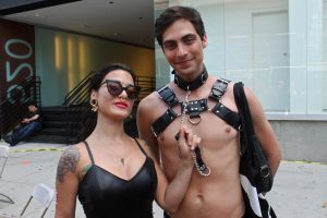 A guy with a leather collar and harness, with a girl holding a chain connected to the collar