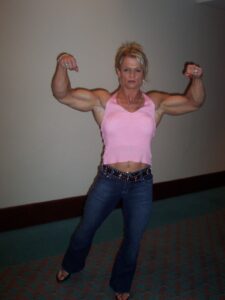 Elena Seiple standing in a hallway doing a double arm bicep flex, wearing a tank top and blue jeans.