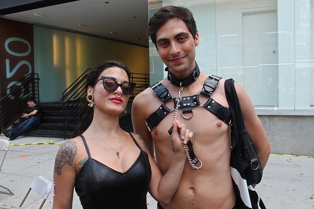 A guy and girl dressed in leather with the girl holding a chain attached to the guy's collar.