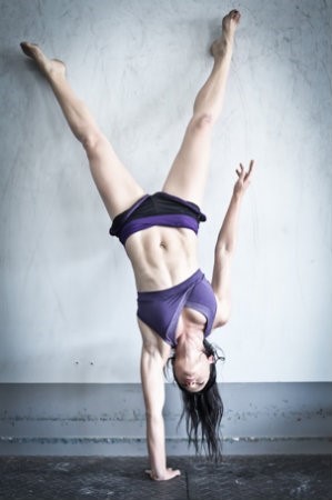 A girl doing a one hand handstand with her feet against the wall.